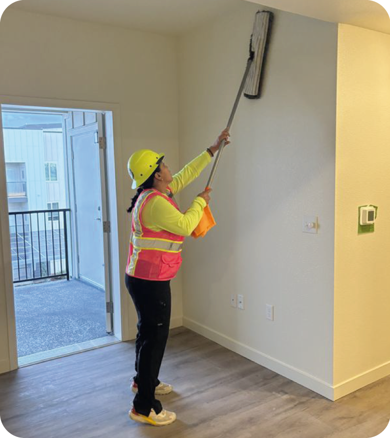 Lola's Shiny Solutions team member in uniform — yellow hard hat, hi-vis vest, and branded shirt — cleaning the upper walls of a newly built apartment unit with an extension mop during a post-construction cleaning job in Tulare County, CA