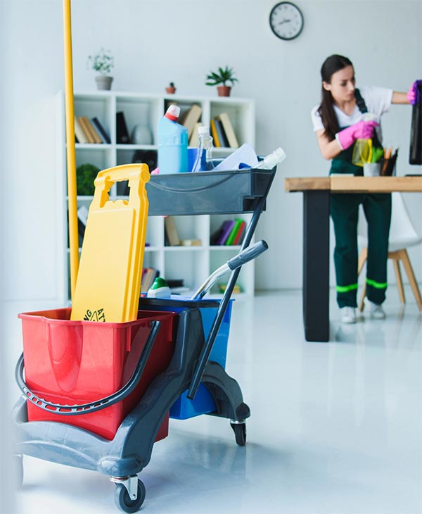 Cleaning cart with mop and buckets in office as staff member cleans desk in background