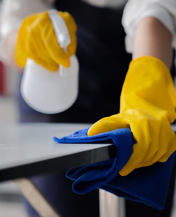 janitorial--about-us-lolas-shiny-solutions Gloved hands wiping down table surface with spray bottle and blue microfiber cloth